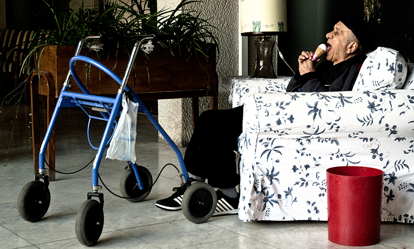 A photo of an old man sitting on a couch eating an ice cream cone with a walker next to him