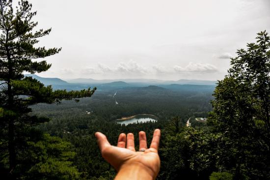 A hand outstretched toward a distant lake from a high vantage point as if presenting that lake.