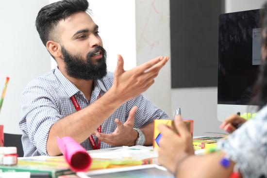 A bearded man sitting at a table gesticulates as if explaining something to a person near him.