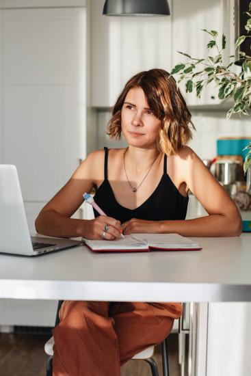 A woman looks into space thoughtfully, poised to write in a journal with laptop open as well.