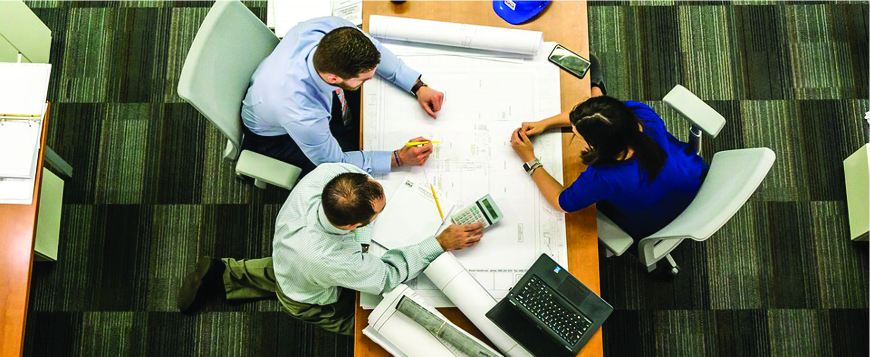 Photograph of three people sitting at a long table with building plans, calculator, and laptop.