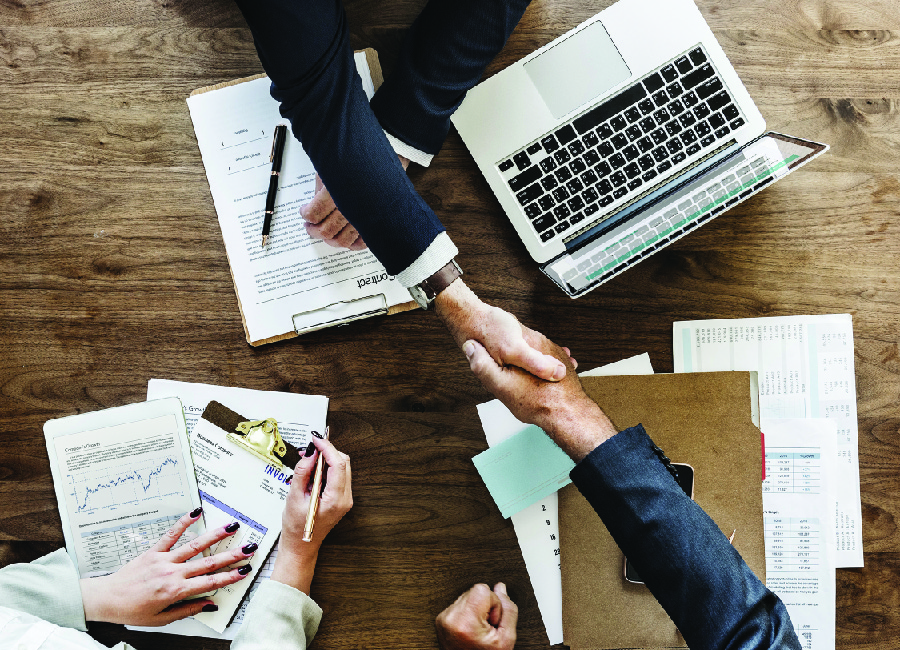 A picture shows two people shaking hands across a table with papers and laptops on it.