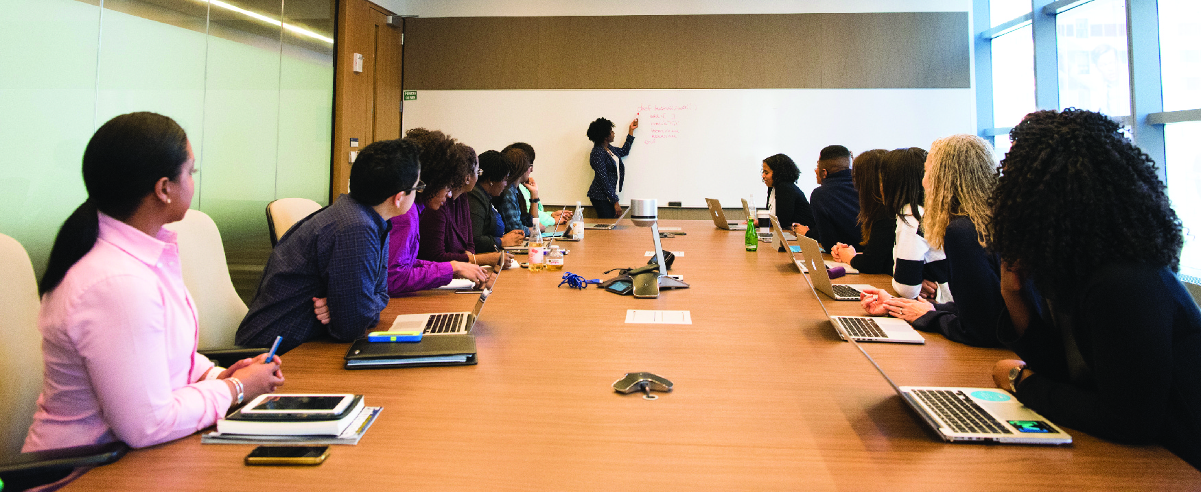 A photograph shows a group of people with laptops and notebooks sitting around a large conference table and looking up at someone writing on a board.